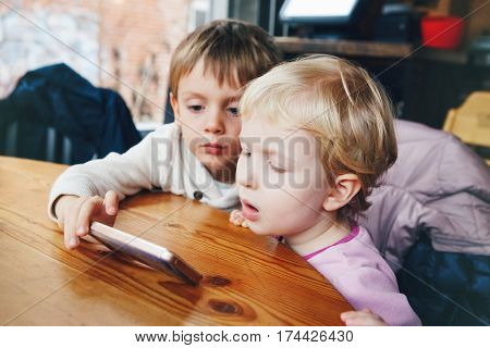 Portrait of two children toddlers boy and girl playing cell phone tablet games watching cartoons movie sitting at table in restaurant real lifestyle candid
