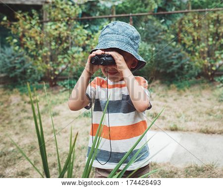 Portrait of cute Caucasian boy in stripped tshirt and hat looking through binoculars outside on house backyard playing spy game candid childhood lifestyle