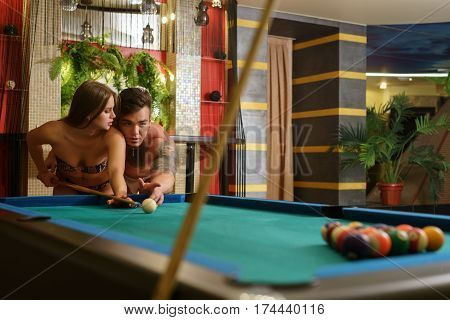 Young handsome athletic tattooed man playing billiards with gorgeous blonde in bicini shot in luxurious sauna interior