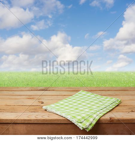 Empty wooden deck table with checked tablecloth over green grass meadow and blue sky background for product montage display