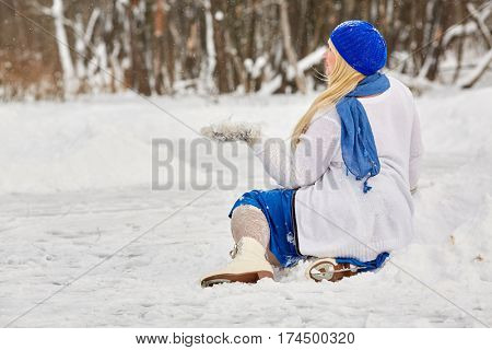Plump smiling woman in skates sits in snowdrift and catches snowflakes on mitten at outdoor skating rink in winter park.