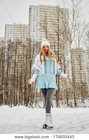 Young woman poses standing on skates at outdoor skate rink in winter park against highrise buildings.