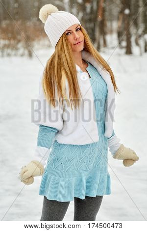 Young woman in blue knitted dress, white fur mantle and hat at outdoor skate rink in winter park.