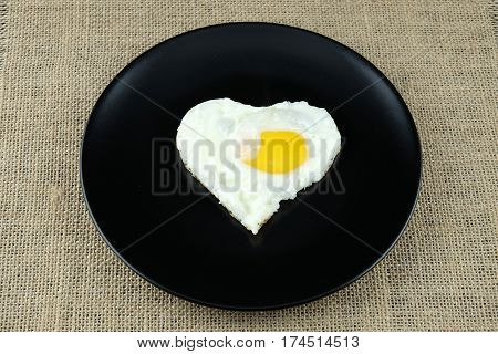 Fried egg in shape of heart on black plate and sackcloth.
