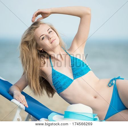 Young lady sunbathing on a beach. Beautiful woman posing at the summer sand beach. Outdoor summer portrait of pretty sport style woman in blue bikini. Ocean sea coast. Beautiful fit tan girl. Sexy slim model caucasian ethnicity outdoors.