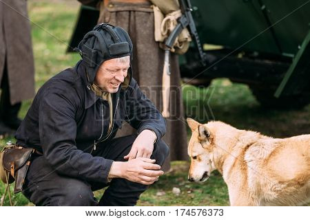 Pribor, Belarus - April 23, 2016: Man Re-enactor Dressed As Russian Soviet Red Army Crew Member Tankman Soldier Of World War II Playing with Dog