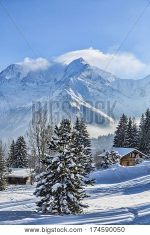 Beautirul winter landscape with wooden chalets and the top of Monta Blanc covered by windy clouds in the background.