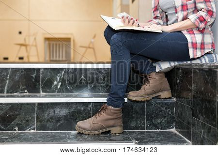Body of female Student in heavy leather Boots jeans Pants and casual Shirt holding notepad and telephone sitting on Stairs in University chat room area