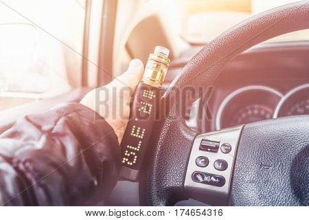 Mans hand on steering wheel of car with modern vaping device or vaporizer, or vape mod, or e-cig gadget. Toned image