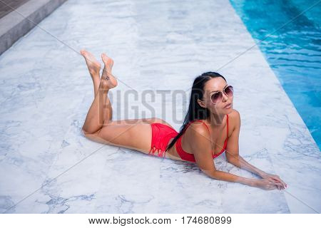 Sexy woman laying and relaxed near pool at cool black fashionable sunglasses, bra bikini pans, tan glowing skin woman, amazing hairstyle, long hair style.