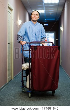 Young man pushing a housekeeping cart laden with clean towels, laundry and cleaning equipment in a hotel as he services the rooms.