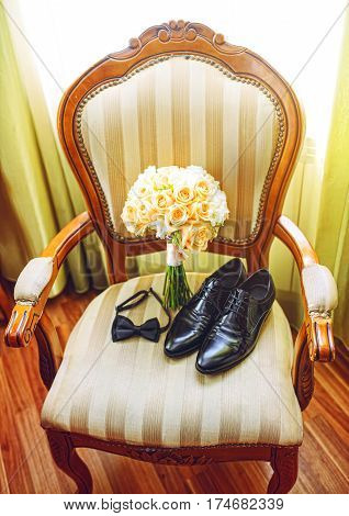wedding bouquet with groom's black shoes and bow tie lying on chair near window.