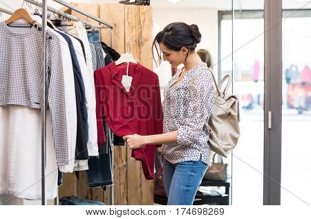 Happy young woman choosing a red blouse from a shop. Young woman in casuals with backpack checking the quality of blouse she is willing to buy. Happy girl buying new clothes for wardrobe.