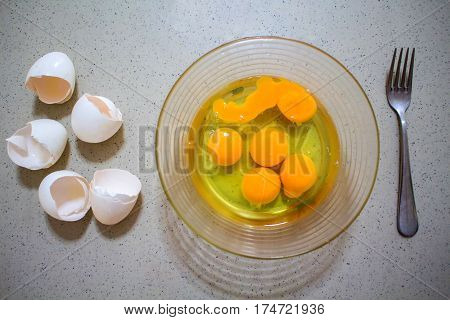 Cooking of eggs in glass bow. Healthy breakfast protein ingredient. Scrambled eggs preparation. Broken eggshells on table. Transparent and yellow content of raw eggs. Nutritional omelette cooking
