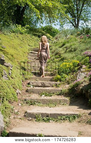 Slim young blonde girl in a brown sundress climbs up the stairs in the park. Sumertime.