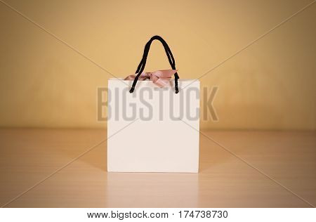 Blank White Paper Gift Bag With A Bow Mock Up Standing On A Wooden Table. Empty Plastic Package Mock