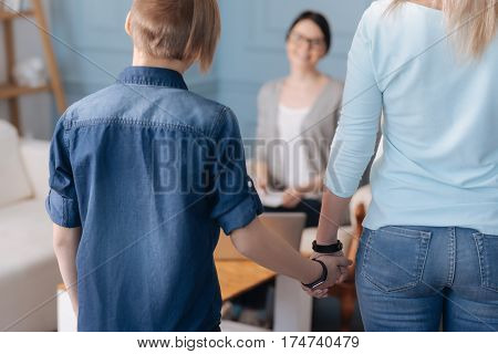 Come to me. Stylish boy wearing jeans shirt taking his mother hand while posing with back on camera