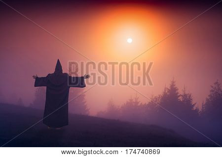 Fairy wizard in a black cassock standing on a hill and welcome raising sun above the foggy valley.