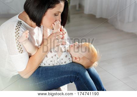 Cute young mother with thick black hair, dressed in a white T-shirt and blue jeans playing with her red-haired young son, dressed in a white shirt and white panties with gray stars, putting it on her lap, sitting on a bed in a light bedroom