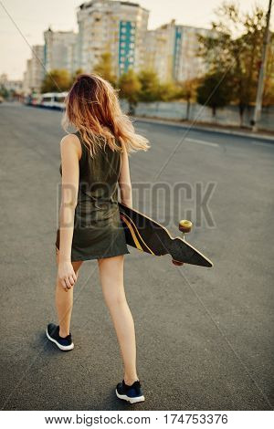 Beautiful young tattooed woman with her longboard on the road in the city in sunny weather