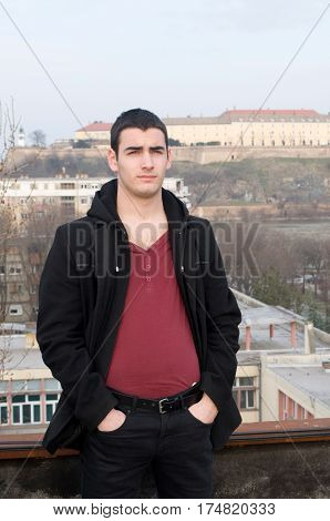 Photographing smiling Danila Mazić in a tight red shirt unbuttoned covered with a black coat and black pants a background Petrovaradin fortress and the Danube River, Novi Sad, Serbia