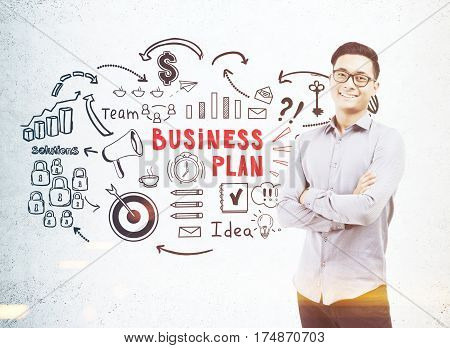 Portrait of an Asian businessman wearing glasses and standing with crossed arms near a concrete wall with a black and red business plan sketch. Toned image