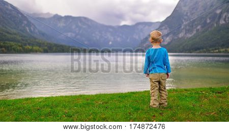 The boy is standing on the green grass on the bank of the lake on a cloudy day