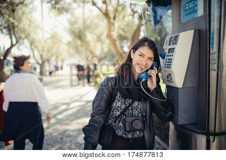 Smiling young woman speaking on a public payphone outside.Happy expression.Dialing local number in foreign country,low cost calls for international dials.Coin or card public phone used by tourist