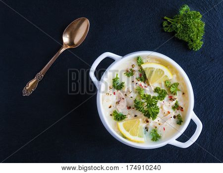 Avgolemono chicken soup with egg-lemon sauce rice and fresh parsley leaves in white bowl on black stone background. Top view.