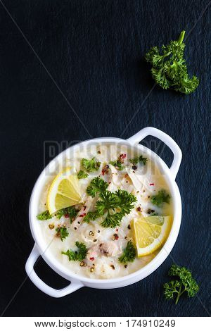Avgolemono chicken soup with egg-lemon sauce rice and fresh parsley leaves in white bowl on black stone background. Top view.