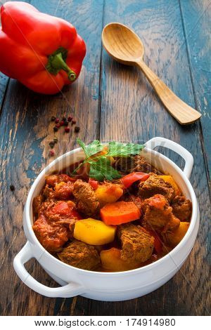 Traditional Hungarian dish goulash slow-cooked with beef potatoes carrot tomatoes paprika onion and bell peppers. White bowl on wooden table.