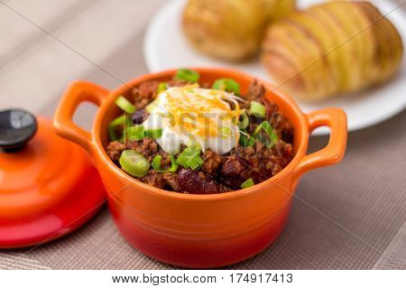 Fresh homemade bowl of chili con carne with beans sour cream grated cheddar cheese and green onions served with hasselback potatoes on the side.