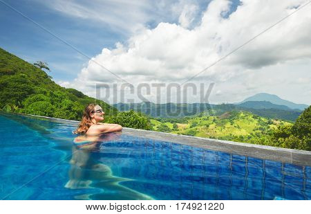 Smile woman enjoying resting on the edge of outdoor swimming pool on background green mountains. Travel happiness emotion summer holiday concept.