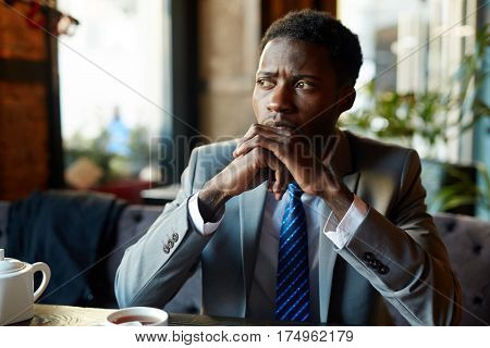 Portrait of handsome African American man wearing business suit looking away to window, pensive and worried while sitting at table in modern restaurant during coffee break