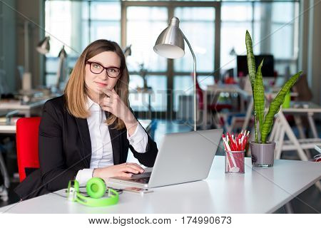 Beautiful Business Lady in official clothing black Jacket and white Shirt sitting at grey Table working on Computer in contemporary digital corporate Office with large Window on Background.