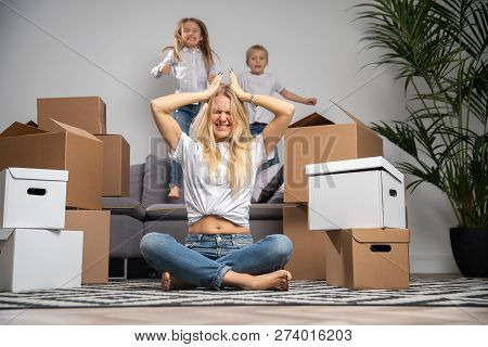Imege Of Displeased Woman Sitting On Floor Among Cardboard Boxes And Boy, Girl Jumping On Sofa In Ne