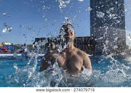 Young Handsome Caucasian Man Splashes In The Pool, Splitting Clear Water, Smiling. Summer And Rest A
