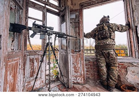 Sniper In A Bulletproof Vest And Helmet Looks Out The Window Near A Large Sniper Rifle