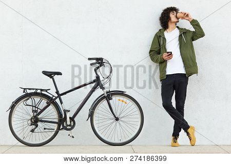 Horizontal Shot Of Fashionable Youngster Keeps Hands Crossed, Holds Mobile Phone, Sends Text Message