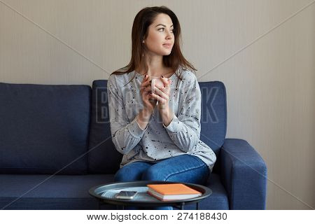 People, Home Concept. Thoughtful Pretty European Woman Holds Cup Of Hot Beverage, Dressed In Sweater