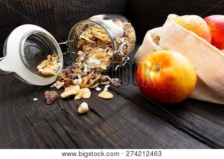 Homemade Granola In Glass Jar And Apples In Reusable Fabric Bag On Dark Wooden Table With Texture. H
