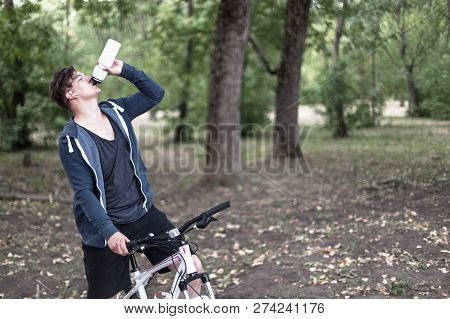 Attractive Young Caucasian Man With Dark Hair Drinks Water From Sport Bin, Bicycling In The Park. Ou