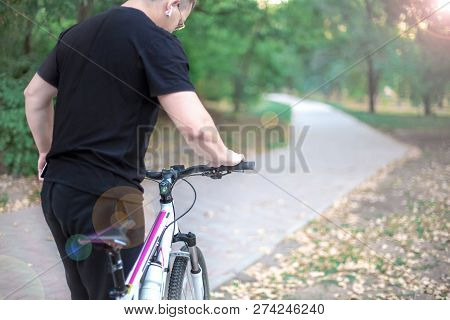 Young Caucasian Man Holds Pink-and-white Bicycle In Front Of Abandoned Path In The Park, Wearing Bla