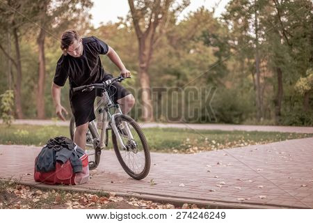 Attractive Young Caucasian Man With Dark Hair On The Bicycle In The Park, Leans Towards His Bag With
