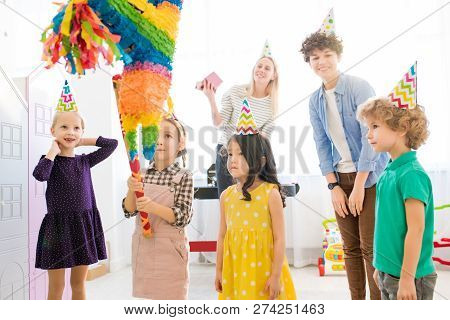 Serious Curious Girl In Denim Suit Holding Bat And Looking At Upside Down House Pinata While Prepari