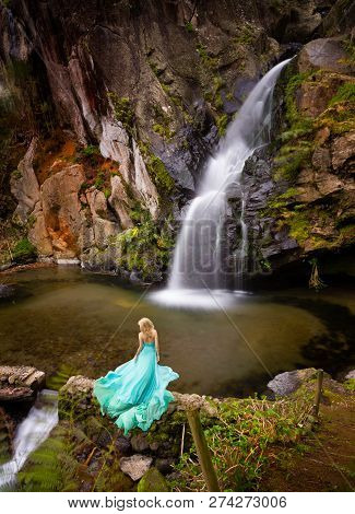 Beautiful Blonde Woman In Elegant Turquoise Long Dress Under A Waterfall Outdoor.