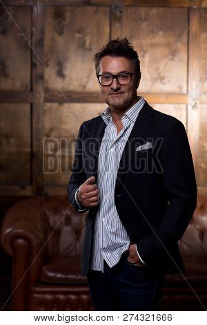 Laconic Portrait Of Positive Middle Aged Man Standing Alone With The Sofa On The Background And Smil