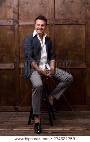 Expressive Dark Haired Man In Stylish Clothes Sitting In The Room Against The Wooden Background And 