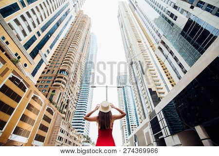 Young Pretty Woman In Red Dress And Straw Hat Walking Beetween Skycrapers In Modern City. Low Angle