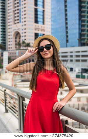 Portrait Of Young Pretty Woman Wearing In Red Dress And Sunglasses, Straw Hat In Front Of Skycrapers